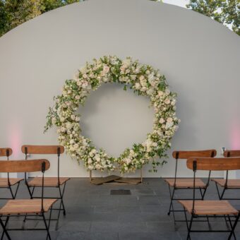 floral circular wedding arch outside on a patio in front of wooden chairs