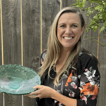 Smiling woman holding a large green glass plate, standing by a wooden fence and leafy plant.