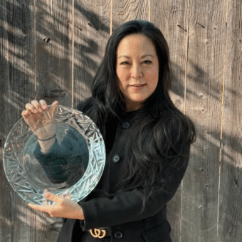 Woman in a black coat holding a large glass plate, standing in front of a wooden fence.
