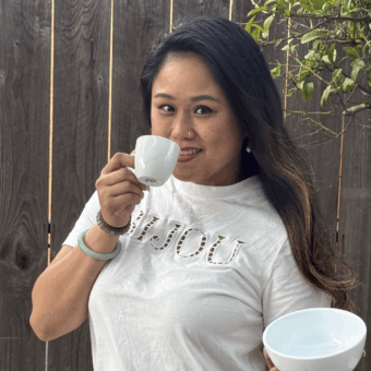 Woman in a white "Bonjour" shirt holding a cup to her mouth and a bowl outdoors by a wooden fence.