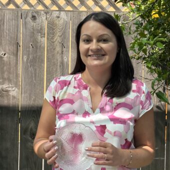 A woman in a pink patterned shirt smiles, holding a clear glass plate outdoors by a wooden fence and greenery.