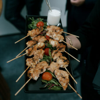 A person holds a tray of grilled chicken skewers with greens, cherry tomatoes, and a small cup of white sauce.