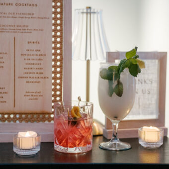 Two cocktails sit on a table with candles and a cocktail menu in the background.